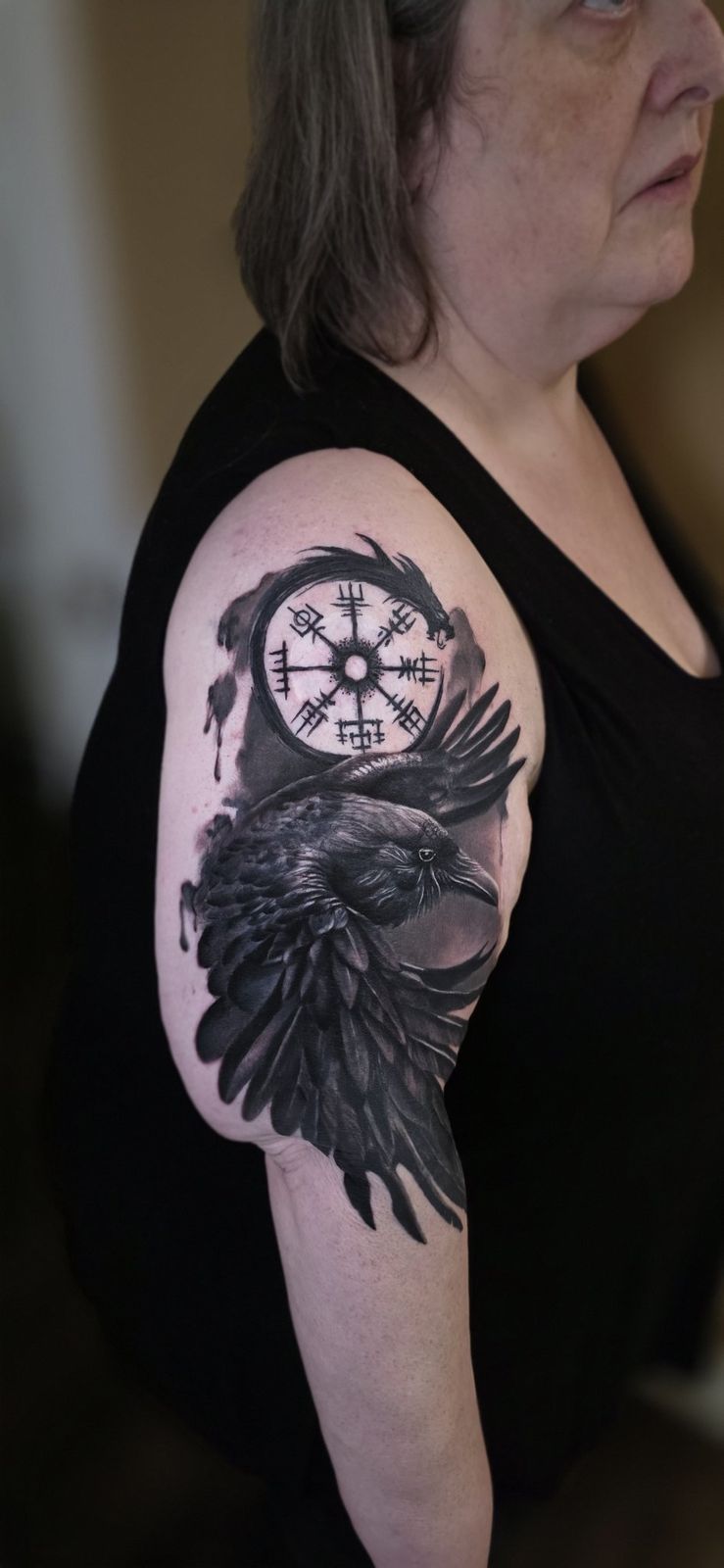 Black-and-gray raven tattoo with a circular symbolic design on the upper arm
