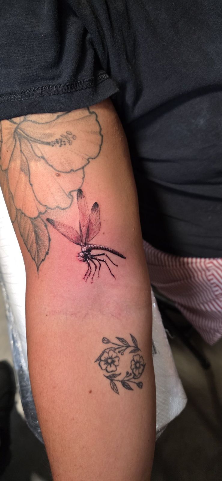 Fine-line dragonfly tattoo shown among existing floral tattoos on the upper arm